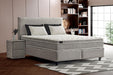 Boxspring double gris clair avec tête de lit rembourrée, matelas à surmatelas intégré et deux oreillers blancs avec coussins noirs, placé dans une chambre moderne avec sol en bois, tapis à motif zigzag et table de chevet assortie